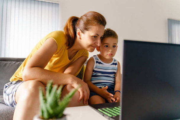 Portré kaukázusi nő és kisfiú ül a laptop előtt otthon - Anya és fia beszél online a közösségi hálón keresztül számítógép segítségével - Valódi emberek kapcsolat szabadidő koncepció - Fotó, kép