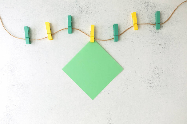 Una hoja verde de papel de escribir cuelga de pinzas de madera de colores en una cuerda, sobre un fondo blanco y maltratado. Puesta plana - Foto, imagen