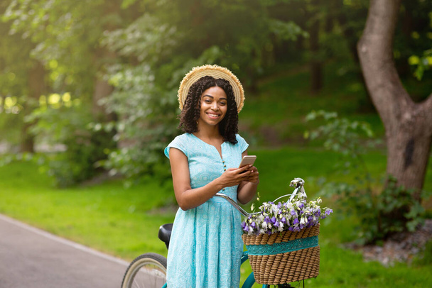 Щаслива чорна дівчина в красивій літній сукні з велосипедом і смартфоном у сільській місцевості
 - Фото, зображення