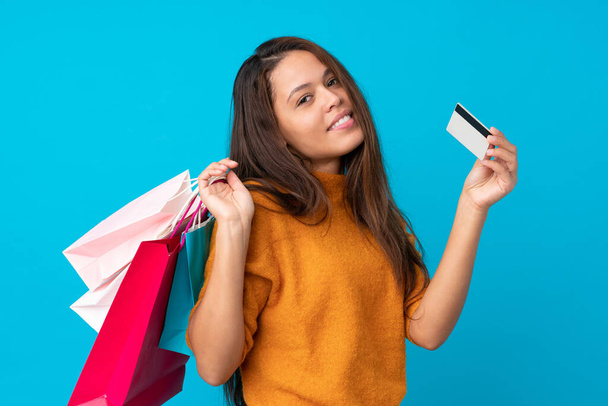 Fiatal brazil lány elszigetelt kék háttér gazdaság bevásárló táskák és egy hitelkártya - Fotó, kép