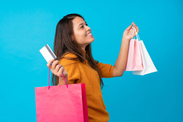 Junge Brasilianerin vor isoliertem blauen Hintergrund mit Einkaufstaschen und Kreditkarte - Foto, Bild