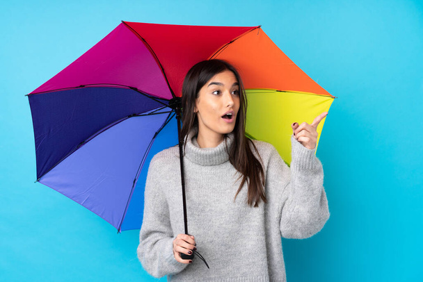 Young brunette woman holding an umbrella over isolated blue wall pointing away - 写真・画像
