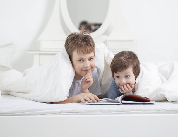 Portrait of a smiling cute brother reading a book lying and relax in bed in a bright white room - Foto, Bild