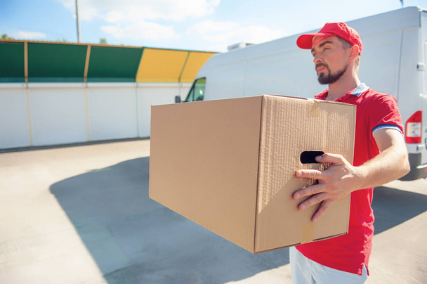 Ein Mann im roten Anzug in der Hand mit einem Karton vor dem Hintergrund des Lieferwagens. Konzept zum Thema Postzustellung - Foto, Bild
