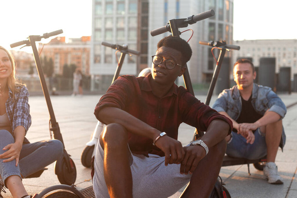 Porträt eines Afroamerikaners in einer europäischen Gruppe von Freunden, junge Leute benutzen Elektroroller und lächeln - Foto, Bild