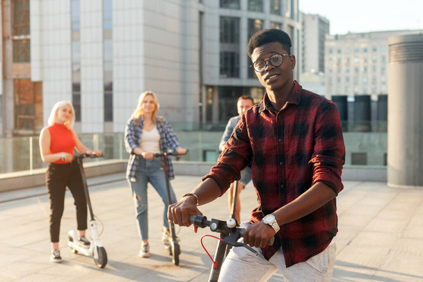afro американец парень с электрическим скутером в компании друзей в городе, межрасовые молодые люди используют экологический транспорт - Фото, изображение
