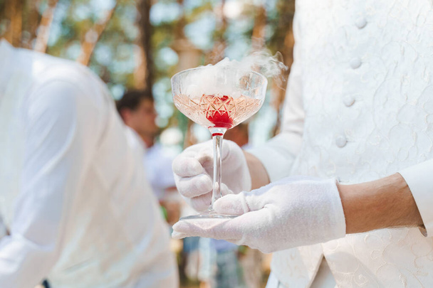Kelner w białych rękawiczkach trzyma kieliszek wina z winem musującym, czerwoną wiśnią i białym dymem z suchego lodu i daje klientowi. Catering na ceremonię ślubną i spotkanie biznesowe. - Zdjęcie, obraz