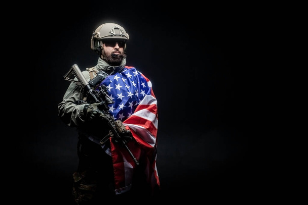 Soldado americano em uniforme militar com uma arma segura a bandeira dos EUA contra um fundo escuro, as tropas de elite da América, forças especiais americanas
 - Foto, Imagem