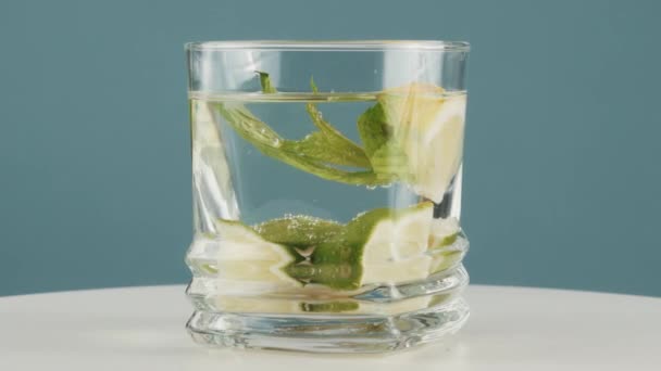 Close up glass of pour water with lime, lemon, mint rotates on blue background - Filmati, video