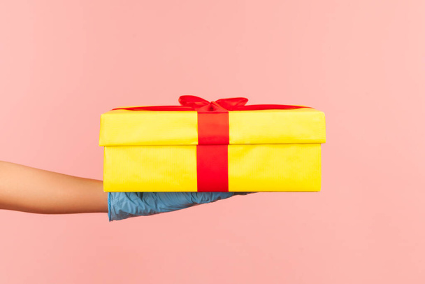 Profile side view closeup of human hand in blue surgical gloves holding yellow gift box. indoor, studio shot, isolated on pink background. - Photo, image