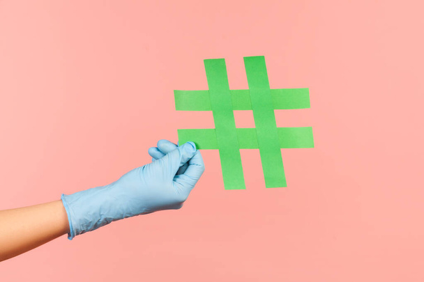 Profile side view closeup of human hand in blue surgical gloves holding green hashtag. indoor, studio shot, isolated on pink background. - Фото, изображение