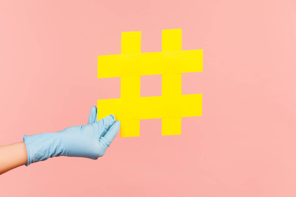 Profile side view closeup of human hand in blue surgical gloves holding yellow hashtag. indoor, studio shot, isolated on pink background. - Φωτογραφία, εικόνα