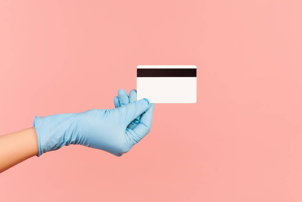 Profile side view closeup of human hand in blue surgical gloves holding classic credit card. indoor, studio shot, isolated on pink background. - Foto, Bild