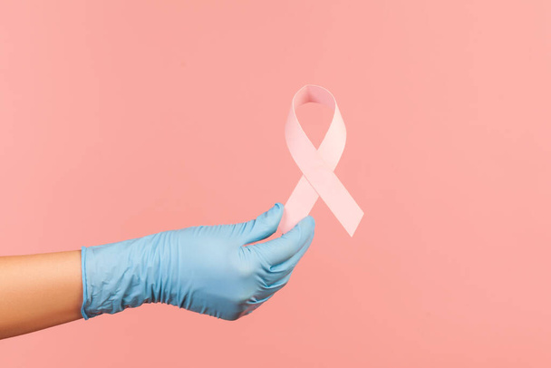 Profile side view closeup of human hand in blue surgical gloves showing and holding pink ribbon, symbol of breast cancer. indoor, studio shot, isolated on pink background. - Photo, image