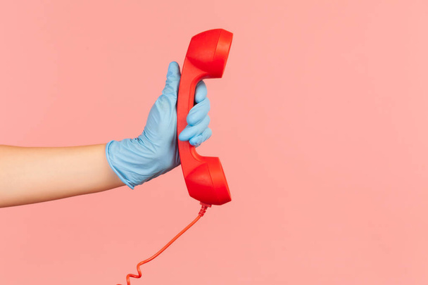 Profile side view closeup of human hand in blue surgical gloves holding and showing red call telephone handset receiver. indoor, studio shot, isolated on pink background. - Valokuva, kuva