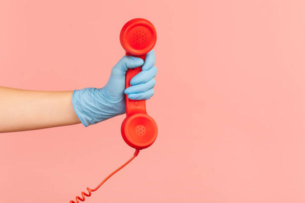 Profile side view closeup of human hand in blue surgical gloves holding and showing red call telephone handset receiver. indoor, studio shot, isolated on pink background. - Фото, изображение