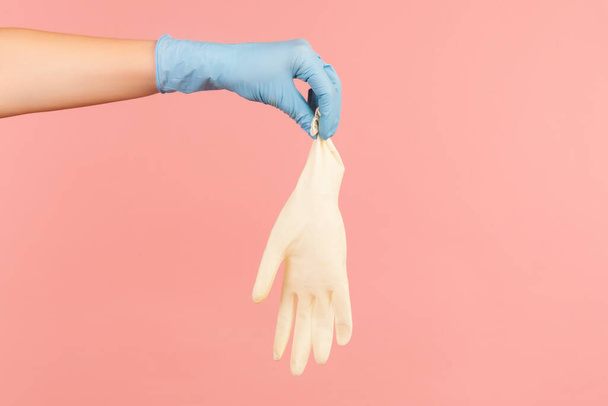 Profile side view closeup of human hand in blue surgical gloves holding white gloves. indoor, studio shot, isolated on pink background. - 写真・画像