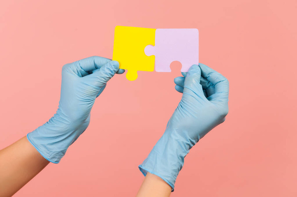 Profile side view closeup of human hand in blue surgical gloves holding yellow and purple puzzle pieses. indoor, studio shot, isolated on pink background. - Zdjęcie, obraz