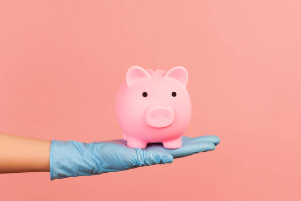 Profile side view closeup of human hand in blue surgical gloves holding pig money box. saving, finicial or banking cocept. indoor, studio shot, isolated on pink background. - Fotó, kép