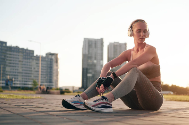 Виснажена спортивна жінка у фітнес-одязі розслабляється під час фізичних вправ і прослуховування музики в навушниках
 - Фото, зображення