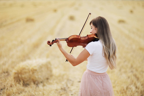 Kaunis romanttinen tyttö löysät hiukset soittaa viulua kentällä sadonkorjuun jälkeen. Neliön lyhteitä pellolla. Viulukoulutus - Valokuva, kuva
