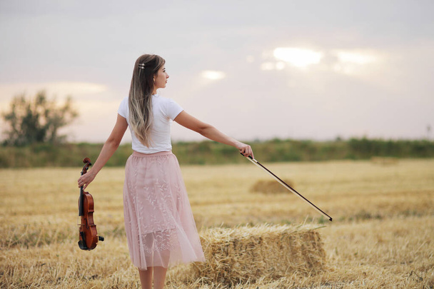 Romantyczna młoda kobieta z falującymi włosami, trzymająca skrzypce w dłoni na polu po zbiorach. Kwadratowe snopki siana na polu. Szkolenie skrzypiec - Zdjęcie, obraz
