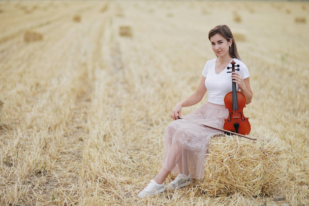 Романтичная молодая женщина с распускающимися волосами, держащая скрипку в руке в поле после сбора урожая. Квадратные снопы сена в поле. Скрипка - Фото, изображение