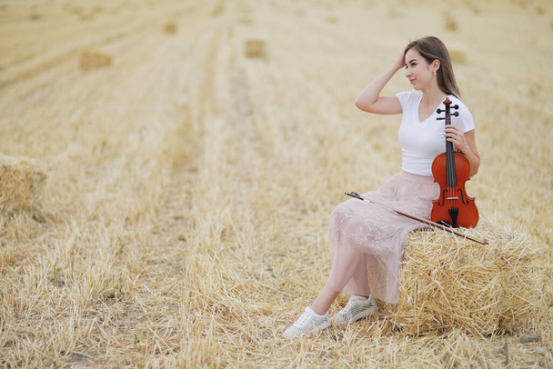 Romantická mladá žena s rozevlátými vlasy, držící po sklizni v ruce housle na poli. Hromádky sena na poli. Trénink houslí - Fotografie, Obrázek