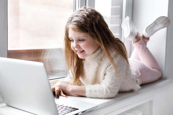 A beautiful girl with a laptop at home at the window looks at the screen and smiles. The girl at home communicates on the Internet. Kids distance learning. - Photo, image