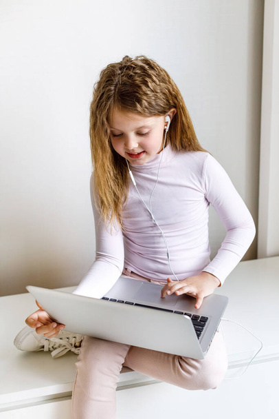 Cute little girl using laptop at home. Vertical photo. The girl at home communicates with friends on the Internet. Education, online study, home studying, schoolgirl children lifestyle concept - 写真・画像