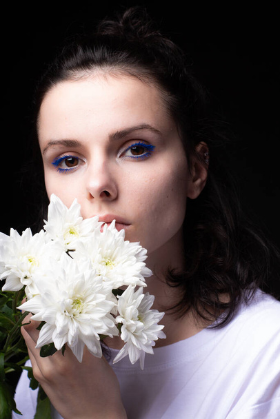 woman with unusual blue makeup holding chrysanthemums in her hands, black studio background - Фото, изображение