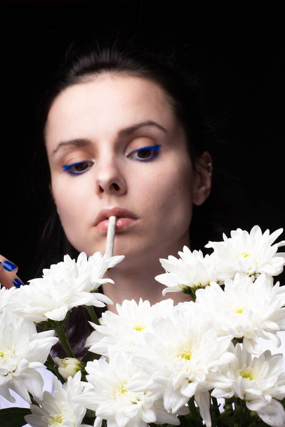 woman with a bouquet of white flowers smokes a cigarette, black studio background - Foto, afbeelding