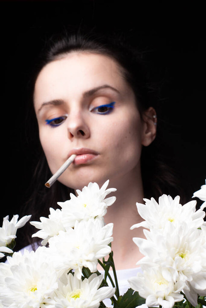 woman with a bouquet of white flowers smokes a cigarette, black studio background - Fotoğraf, Görsel