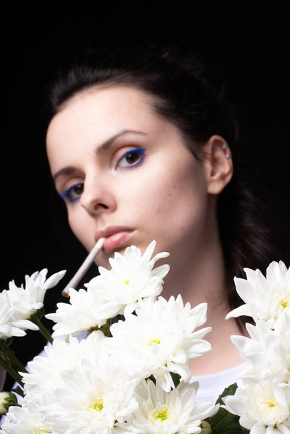 woman with a bouquet of white flowers smokes a cigarette, black studio background - Photo, Image