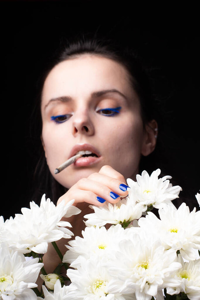 woman with a bouquet of white flowers smokes a cigarette, black studio background - Foto, Imagem