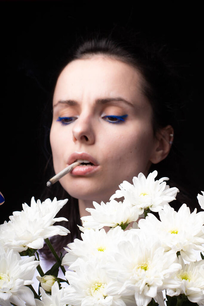 donna con un mazzo di fiori bianchi fuma una sigaretta, sfondo nero studio - Foto, immagini