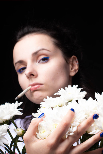 woman with a bouquet of white flowers smokes a cigarette, black studio background - Zdjęcie, obraz