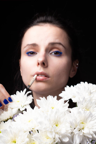 woman with a bouquet of white chrysanthemums smokes a cigarette, dark studio background - Foto, afbeelding