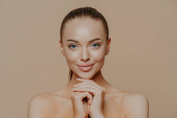 Young pretty woman with beauty skin and combed hair, keeps hands under chin, has healthy complexion after face purification, soft well cared body, isolated over studio wall. Facial treatment - Photo, Image