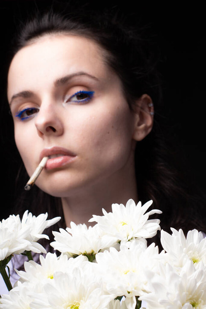 woman with a bouquet of white chrysanthemums smokes a cigarette, dark studio background - Фото, зображення
