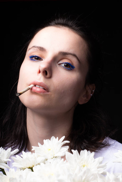 woman with a bouquet of white chrysanthemums smokes a cigarette, dark studio background - Фото, изображение