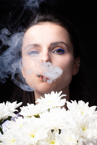 femme avec un bouquet de chrysanthèmes blancs fume une cigarette, fond sombre studio - Photo, image