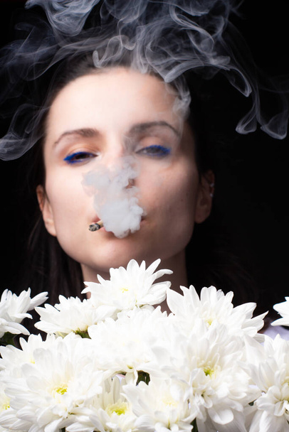 Frau mit einem Strauß weißer Chrysanthemen raucht eine Zigarette, dunkler Studiohintergrund - Foto, Bild