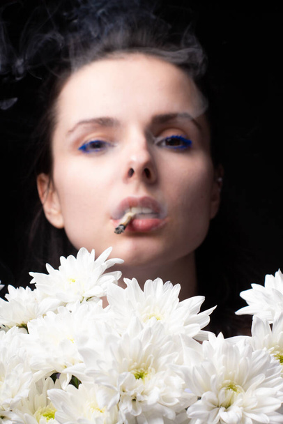 woman with a bouquet of white chrysanthemums smokes a cigarette, dark studio background - Фото, зображення