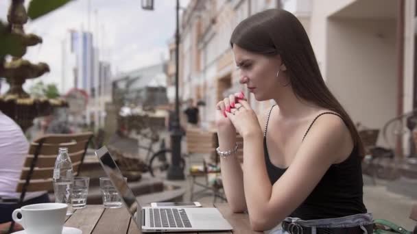 Een leuke studente zit aan een tafel in een café op straat. Ze leest iets smerigs en slaat de laptop. Moderne technologie en jeugdconcept. - Video