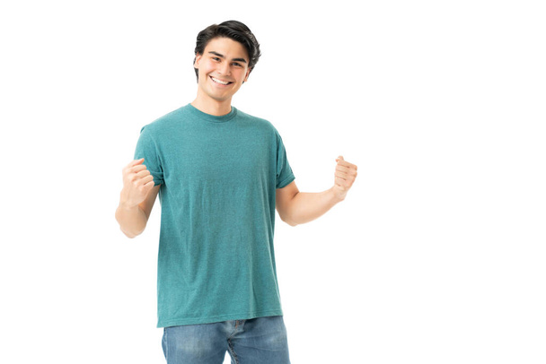 Lächelnder hispanischer junger Mann mit geballten Fäusten feiert Sieg im Studio mit weißem Hintergrund - Foto, Bild