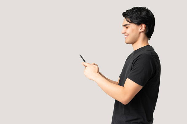 灰色の背景を持つスタジオに立っている間、スマートフォンを通して笑顔若い男のテキストメッセージの側面図 - 写真・画像