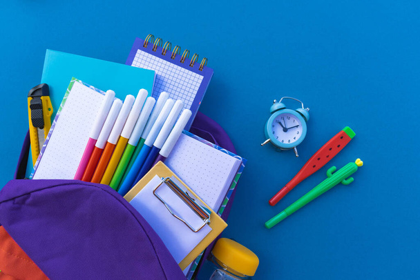 Stationery for children are falling out of a backpack. Blue background. Kawaii office supplies. Bright trend color Flat lay Copy space - Foto, Imagen