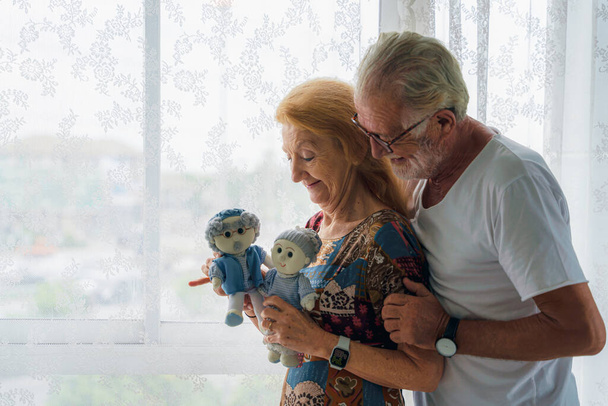 caucasian senior husband and wife couple standing by windowat home with old dolls on hand showing happy and romantic face of loving each other - Foto, Imagen