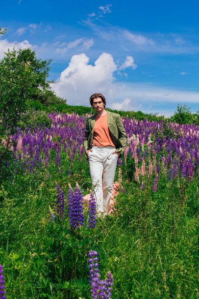 Tall handsome man in a green jacket walking on a pink cloth in lupine flowers field and enjoing the beauty of nature. Man surrounded by purple and pink lupines. - Foto, Imagem